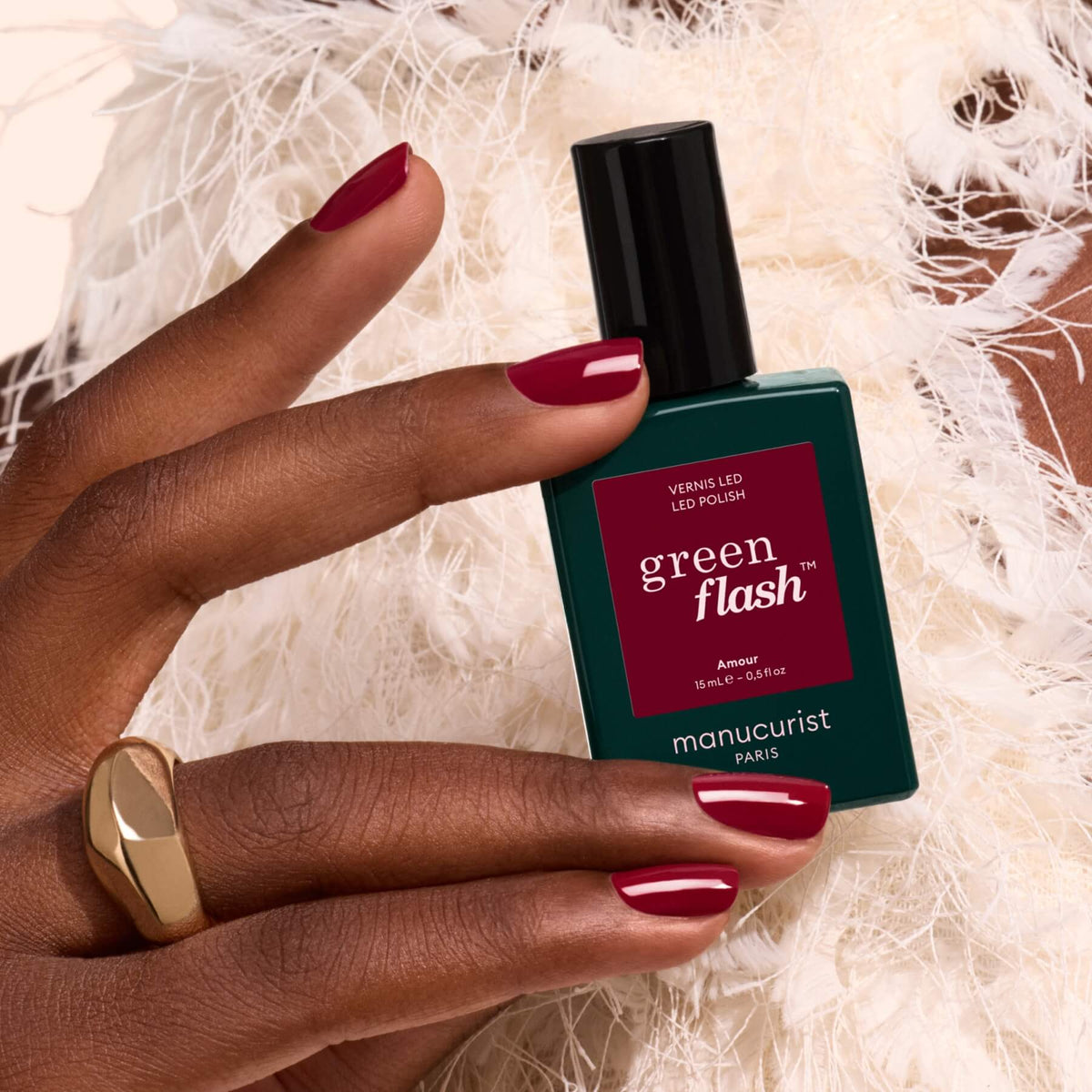 Une main aux ongles vernis d’un bordeaux lumineux tient un flacon vert de vernis gel Amour de Manucurist, avec des plumes blanches en arrière-plan.