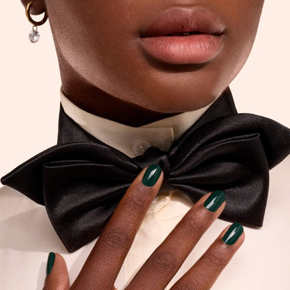 A person with dark skin wears a black bow tie, cream shirt, and gold earring. Their nails, painted in Manucurist’s Emerald shade, gently touch the bow tie.
