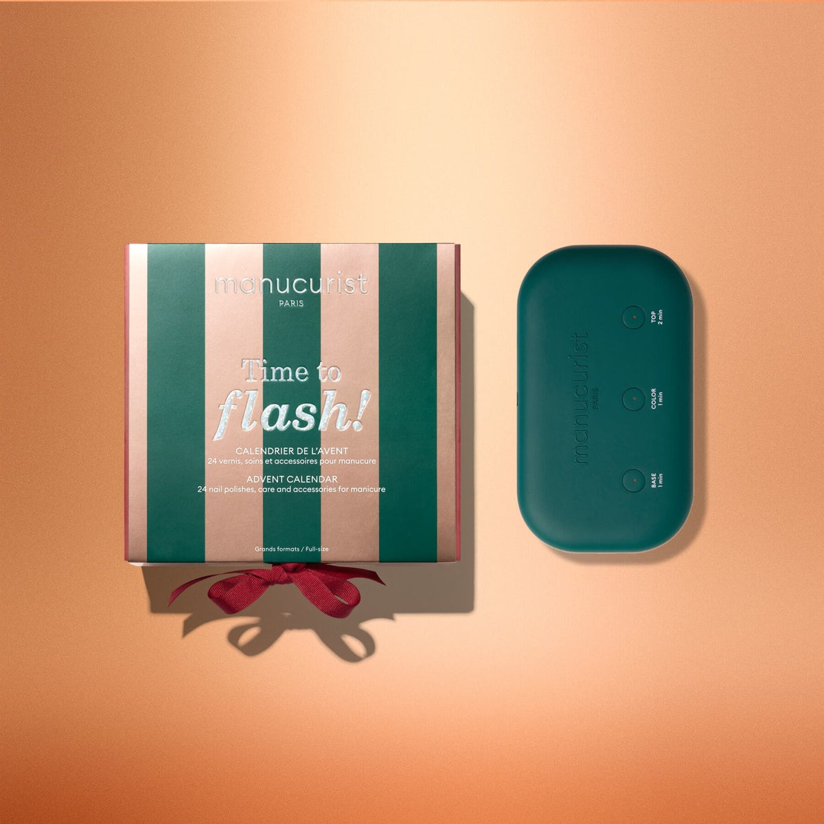 A green and pink striped Manucurist Advent Calendar Time to Flash! box, inspired by the Green Flash gel polish manicure and decorated with a red bow at the bottom, sits next to a teal manicure UV lamp on a peach-colored background.