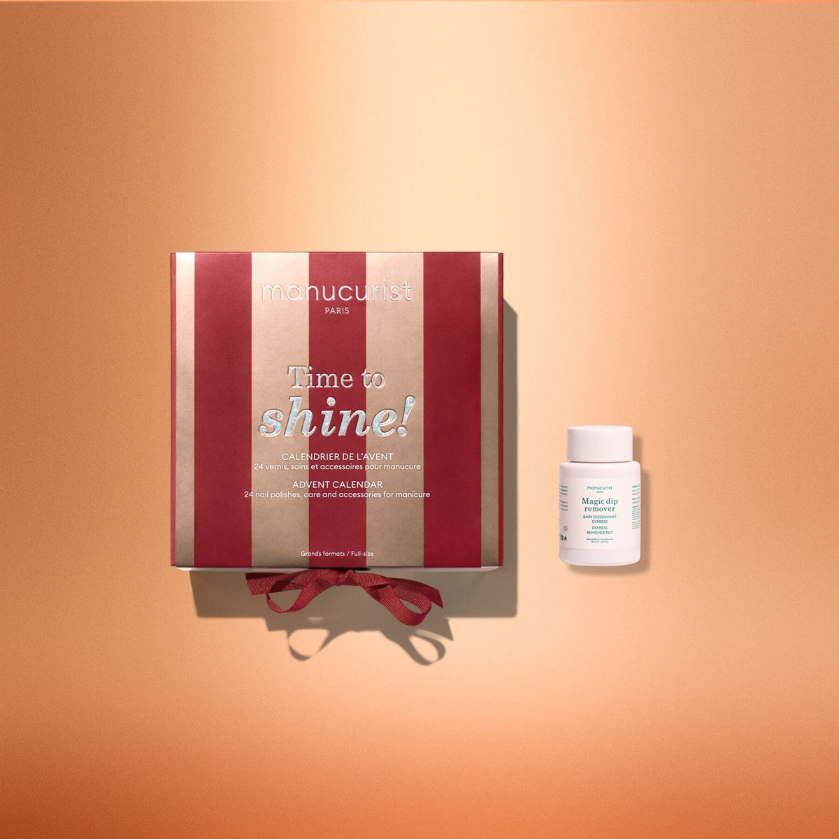 A red and gold striped box labeled “Manucurist Advent Calendar Time to Shine!” rests beside a small white bottle on a peach gradient background. This festive Advent Calendar from Manucurist contains premium nail and hand care essentials, including the Active™ Shine Icy treatment for dazzling results.