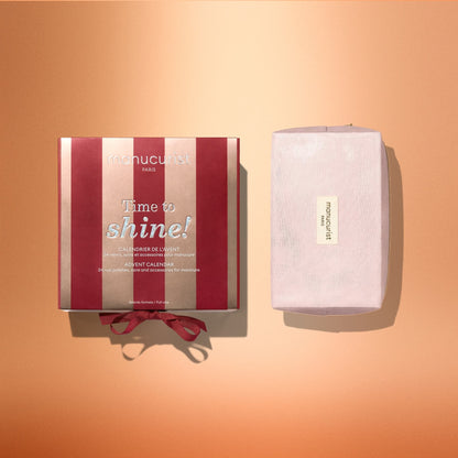 A red and pink striped Manucurist Advent Calendar Time to Shine! box for nail and hand care, featuring “Time to shine!” on the front, sits beside a pink-wrapped rectangular gift on a peach background, hinting at the surprise of discovering Active™ Shine Icy treats inside.