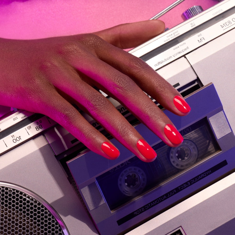 A hand with glossy Spicy red nail polish by Manucurist rests on a silver retro cassette tape player, illuminated by vibrant pink lighting and subtle undertones.