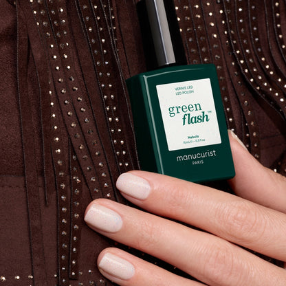 A hand with neatly manicured, milky white gel nails holds a bottle of Manucurist Nebula polish against a brown, beaded, fringed fabric background.