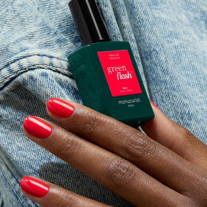 A hand with glossy pop red "Spicy" gel polish nails holds a bottle of Manucurist Spicy against a light blue denim background.