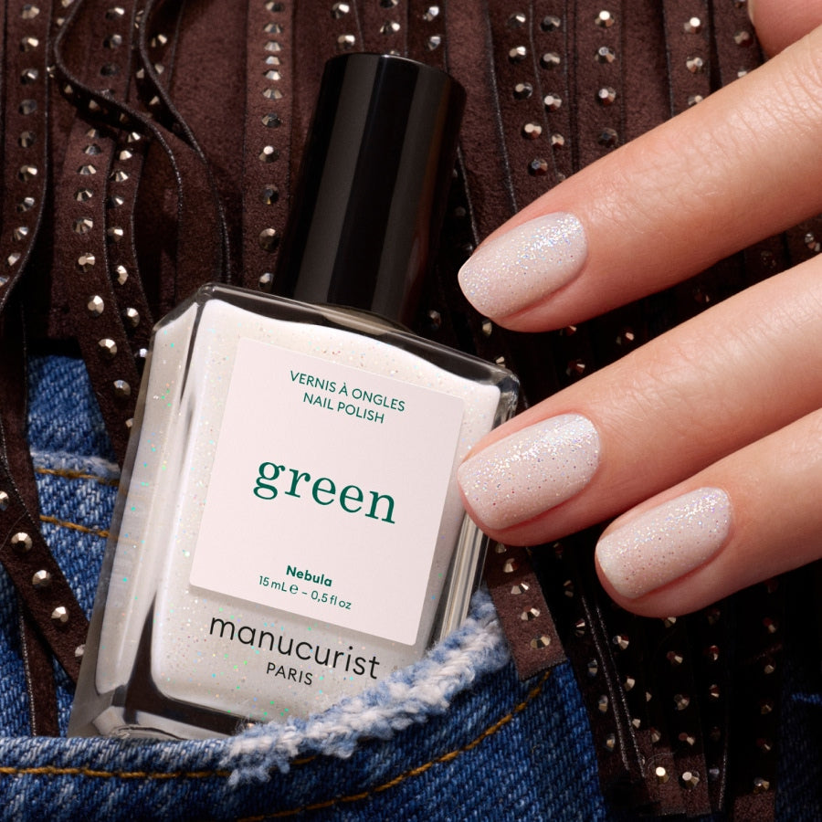 A hand with short, shimmering pale nails holds a bottle of Manucurist Nebula polish against blue denim and brown fabric with metal studs.