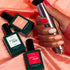 A hand with red and peach nails holds a silver lamp over pink velvet beside three Manucurist Curated Trios bottles—Green Flash polish in green and red—ideal for chic, modern manicures.
