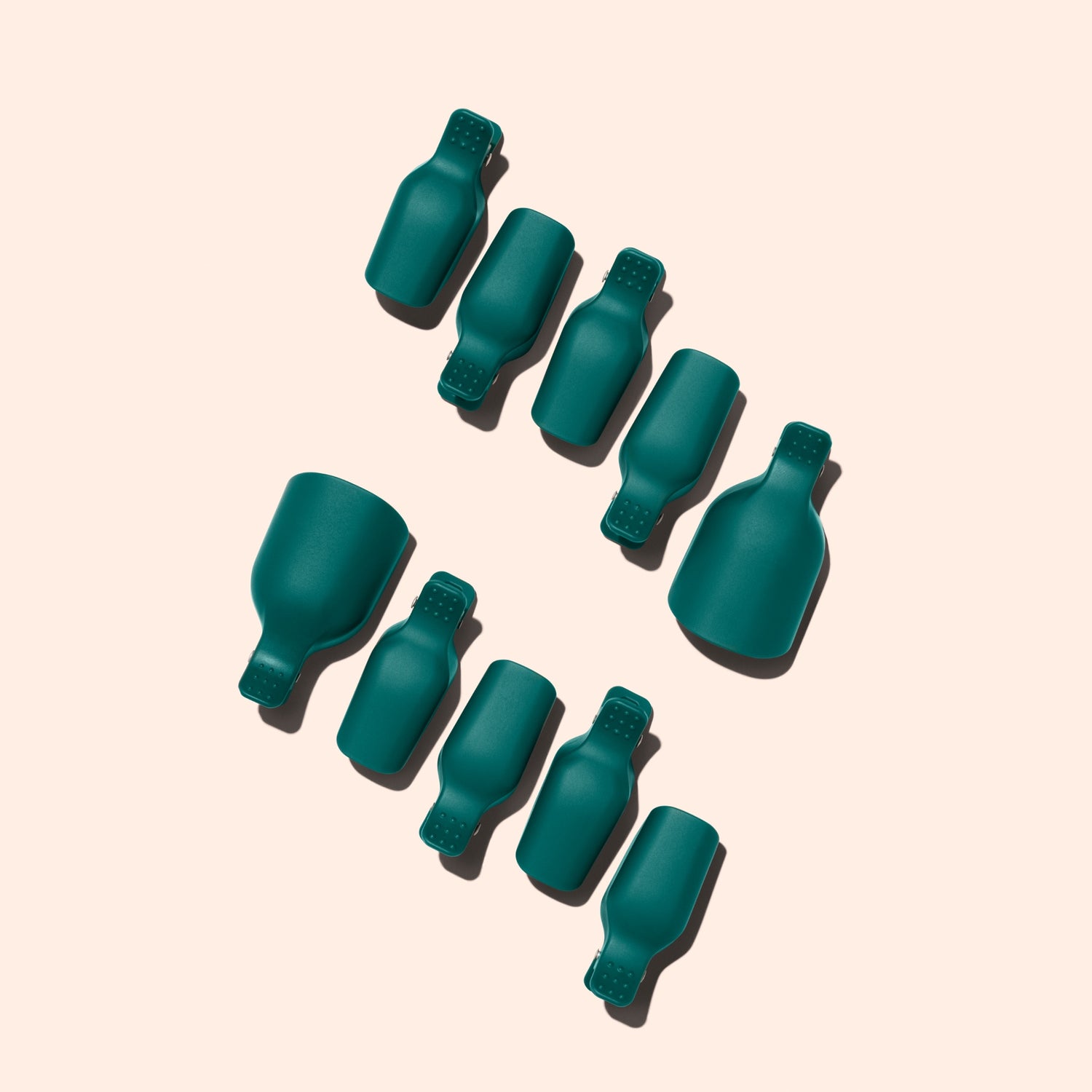 Nine teal Foot Removal Clips by Manucurist, shaped like bottles, are arranged diagonally on a light beige background. Each casts a shadow to the left, with two clips noticeably larger than the others.
