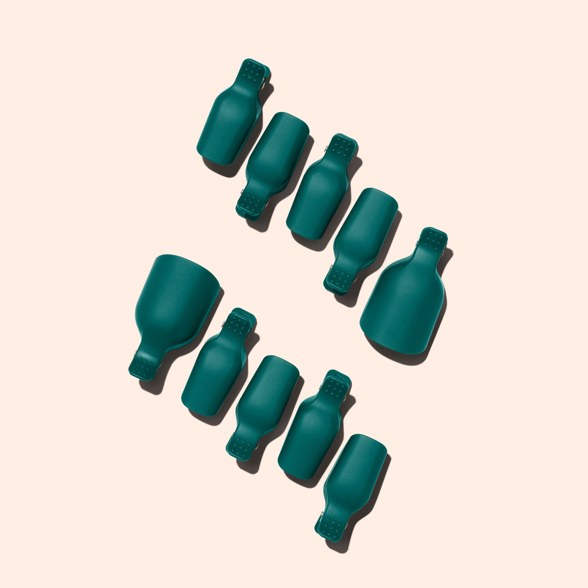 Neuf Foot Removal Clips bleu sarcelle de Manucurist, en forme de flacons, sont disposés en diagonale sur un fond beige clair. Chacun projette une ombre vers la gauche, deux clips étant nettement plus grands que les autres.