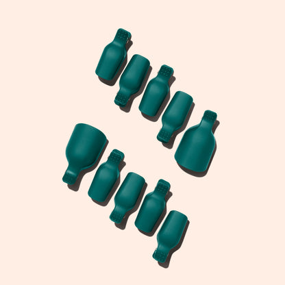 Nine teal Foot Removal Clips by Manucurist, shaped like bottles, are arranged diagonally on a light beige background. Each casts a shadow to the left, with two clips noticeably larger than the others.