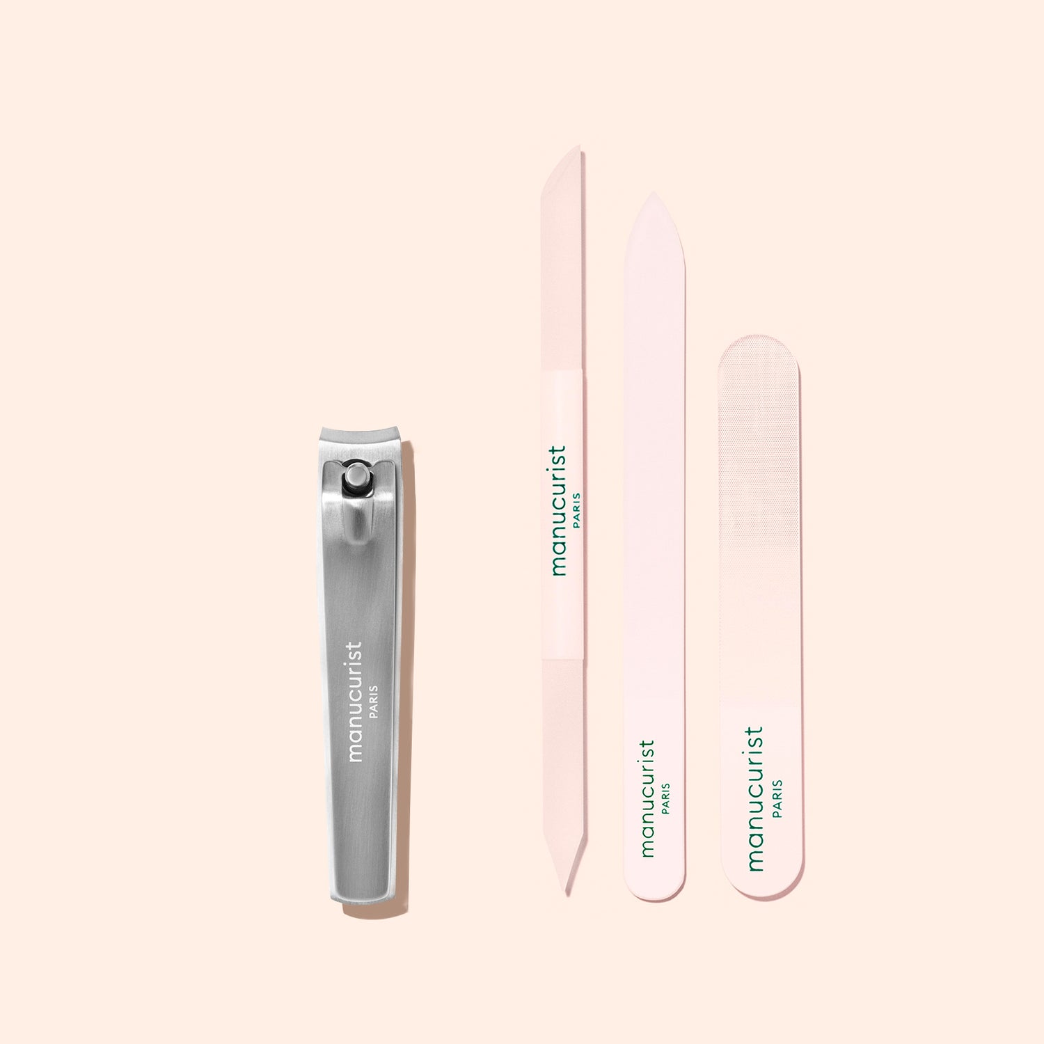 A Manucurist Nail Clipper in silver and three pastel pink nail files, each bearing the Manucurist brand, are arranged neatly on a light beige background.