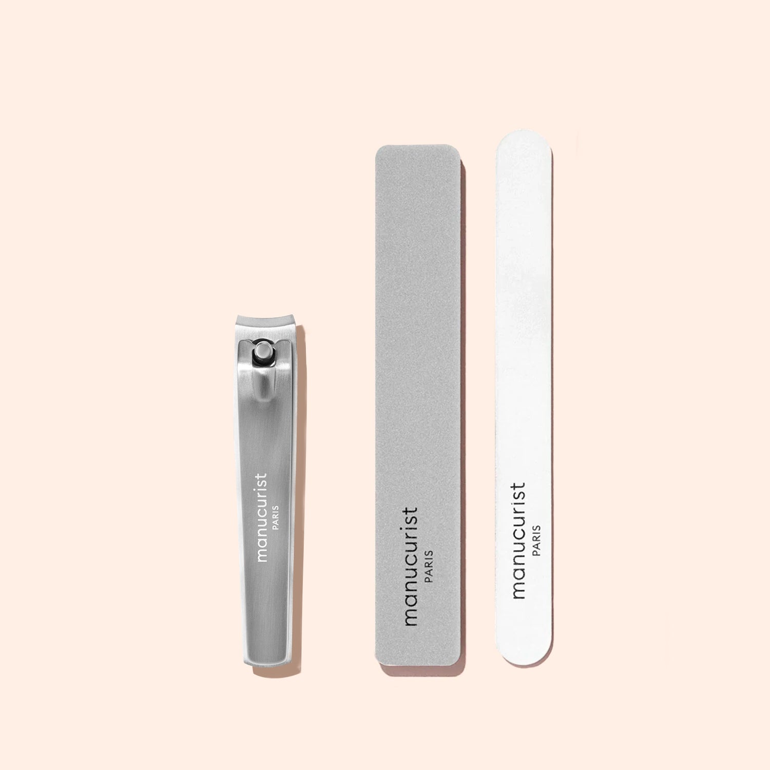 A Manucurist Nail Clipper, accompanied by a grey and a white nail file, are arranged vertically on a light beige background. All items feature the Manucurist brand name.