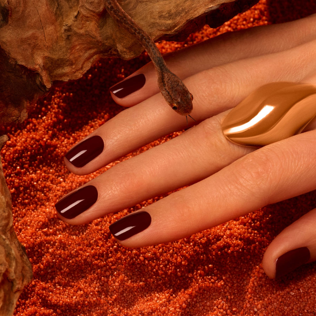 A hand with Grenat, a rich earthy red polish by Manucurist, rests on reddish sand. A small snake slithers over the fingers, which are adorned with a large wavy gold ring.