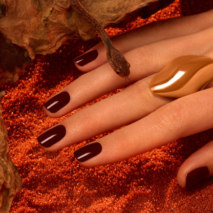 A hand with Grenat, a rich earthy red polish by Manucurist, rests on reddish sand. A small snake slithers over the fingers, which are adorned with a large wavy gold ring.