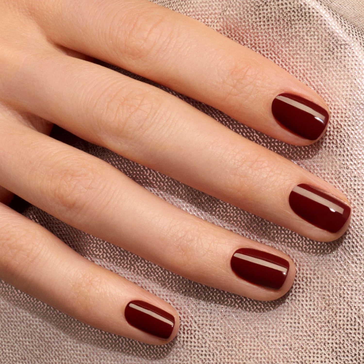 A close-up of a hand with neatly manicured nails painted in Manucurist’s Grenat, a glossy maroon red gel polish, resting on a light textured fabric.