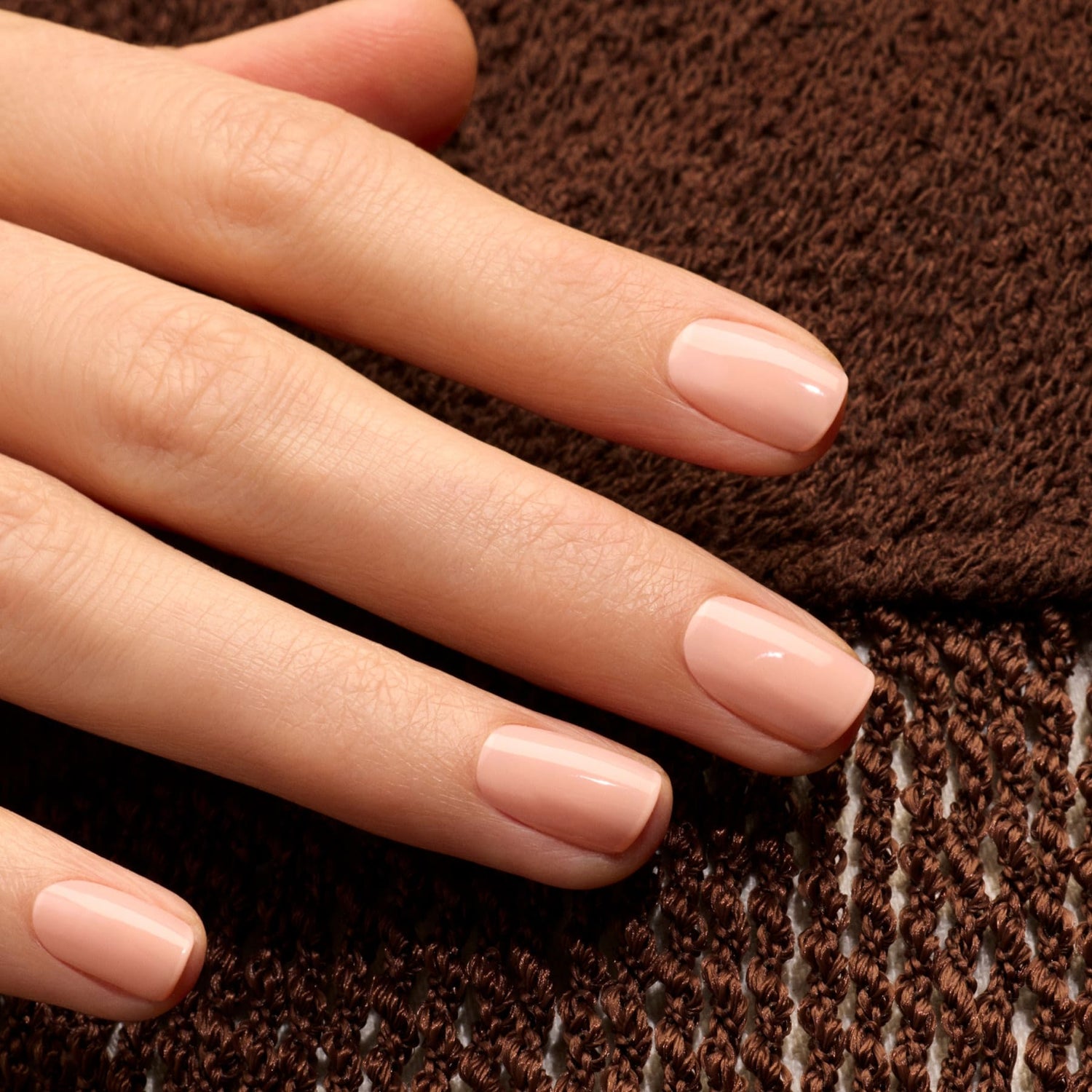 A close-up of a hand with neatly manicured nails painted in Manucurist’s Milky Peach gel polish, resting on brown textured fabric.