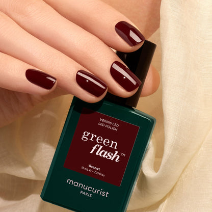 A hand with short, earthy red polished nails holds a bottle of Manucurist Green Flash gel polish in the shade Grenat against a light, soft fabric background.