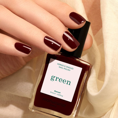 A hand with neatly manicured, maroon red nails holds a bottle of Manucurist Grenat nail polish. The background features a soft, cream-colored fabric.