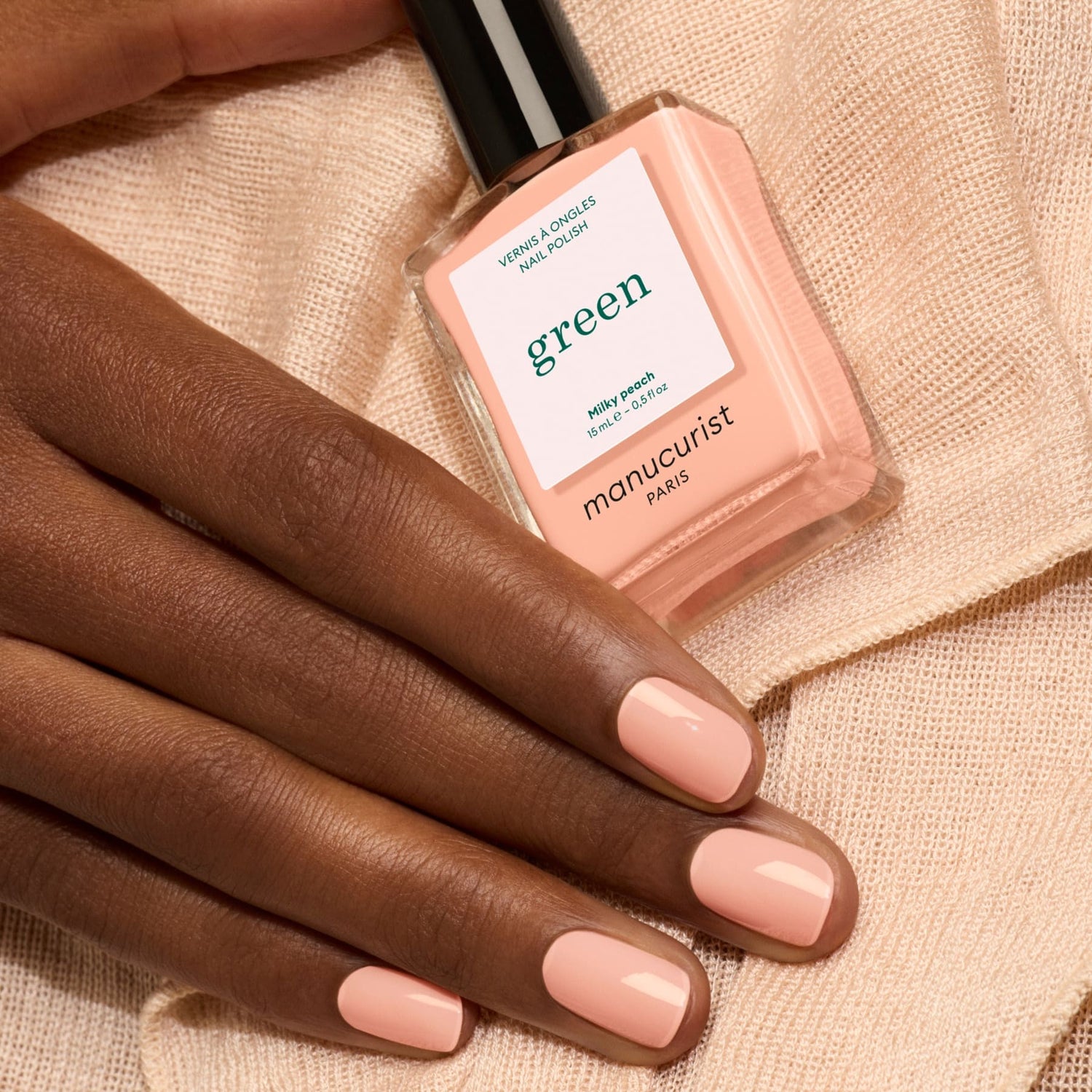A hand with neatly manicured nails painted a soft, milky peach rests on gentle beige fabric. Nearby, a bottle of Manucurist Milky Peach nail polish completes the elegant scene.