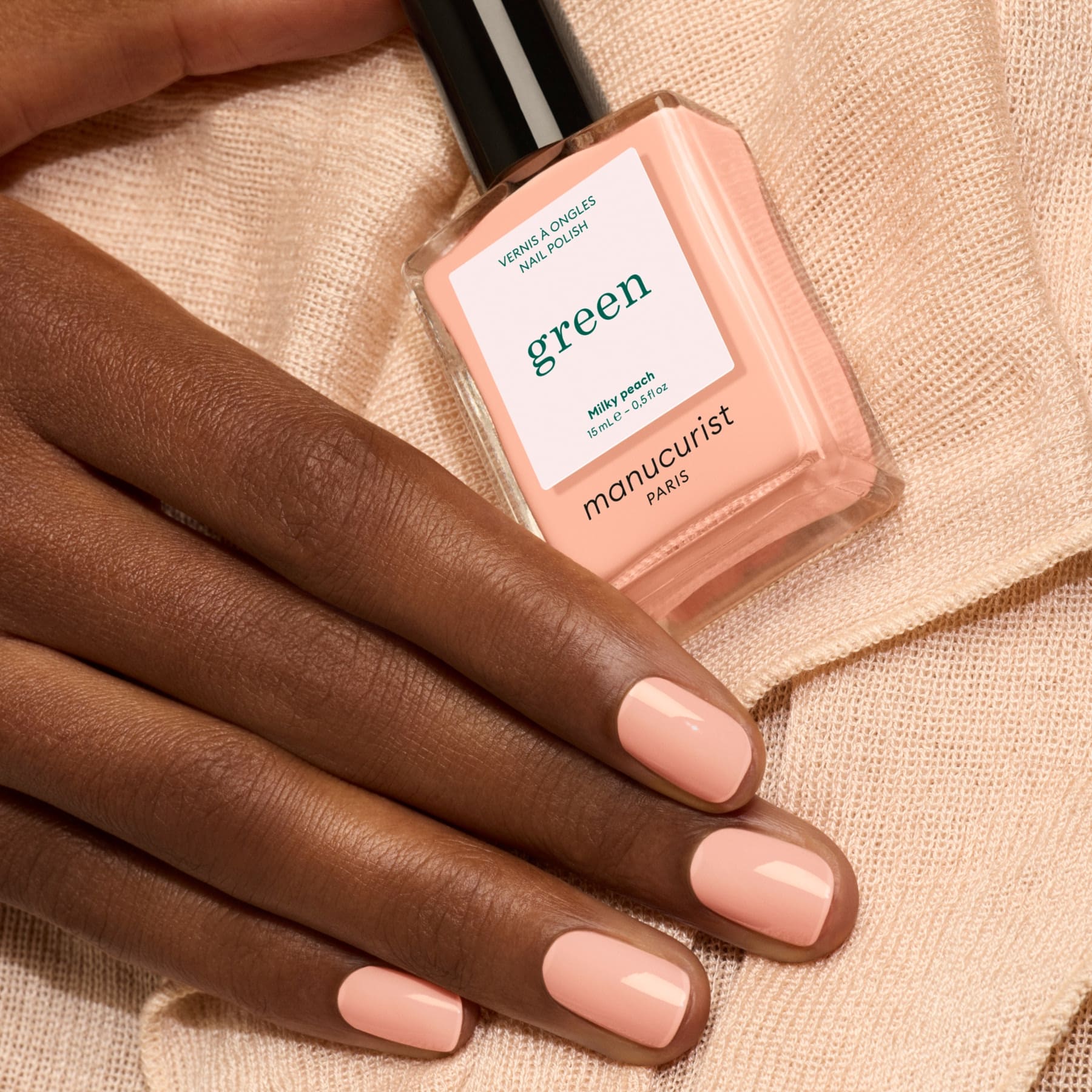 A hand with neatly manicured nails painted a soft, milky peach rests on gentle beige fabric. Nearby, a bottle of Manucurist Milky Peach nail polish completes the elegant scene.