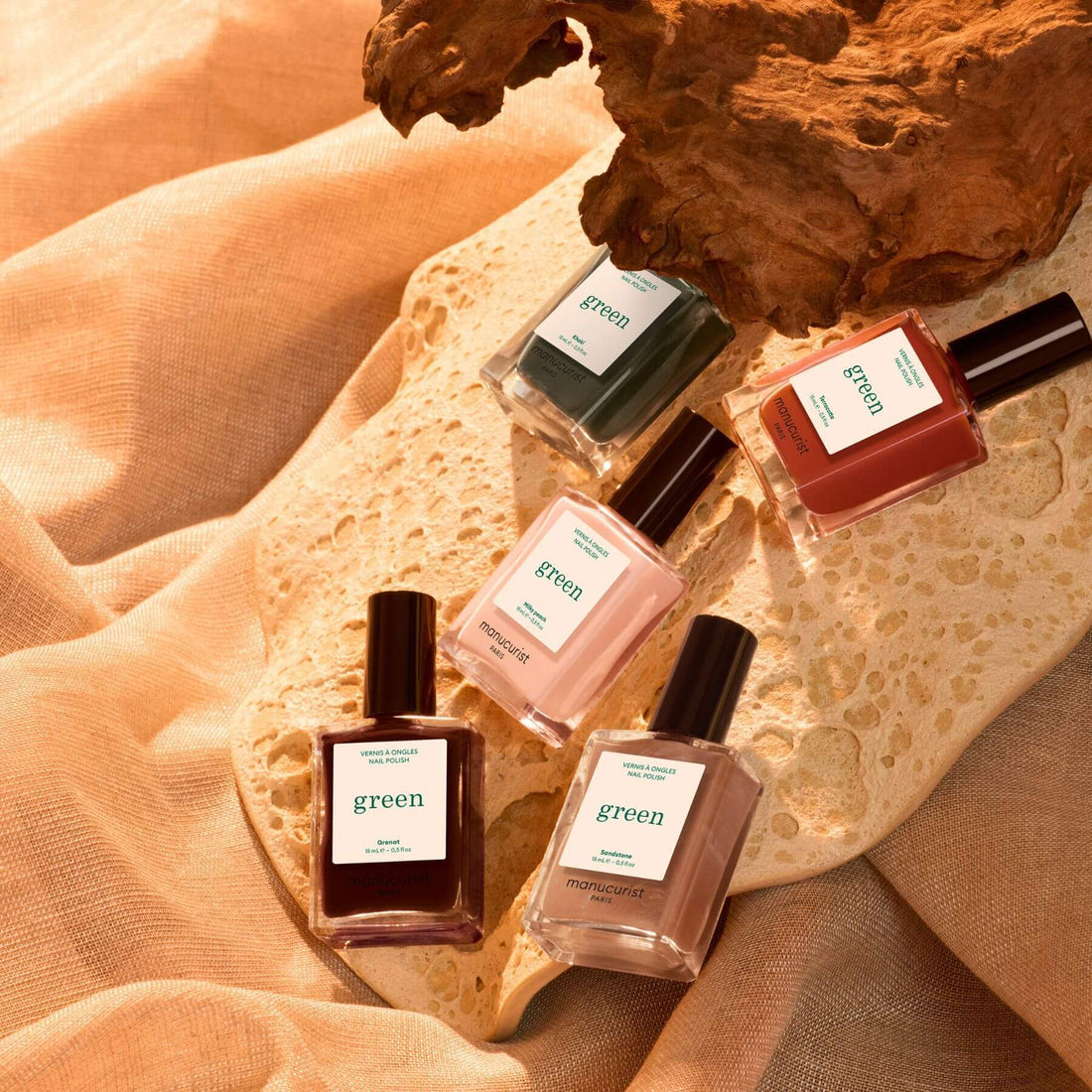 Four bottles from Manucurist’s Curated Color Collections rest on a textured beige surface, complemented by natural elements like stone and wood, with warm lighting enhancing their earthy hues—offering perfect inspiration for the latest nail trends and thoughtfully curated palettes for your next manicure.