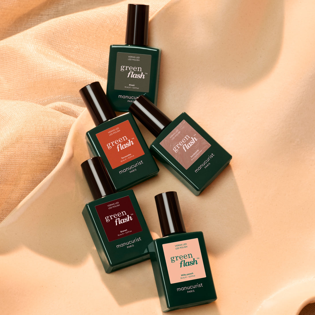 Five bottles from the Manucurist Curated Color Collections are arranged diagonally on a softly textured beige fabric background. Each features the brand’s signature green cap and distinctively colored labels, artfully highlighting inspiring palettes perfect for creative manicures.
