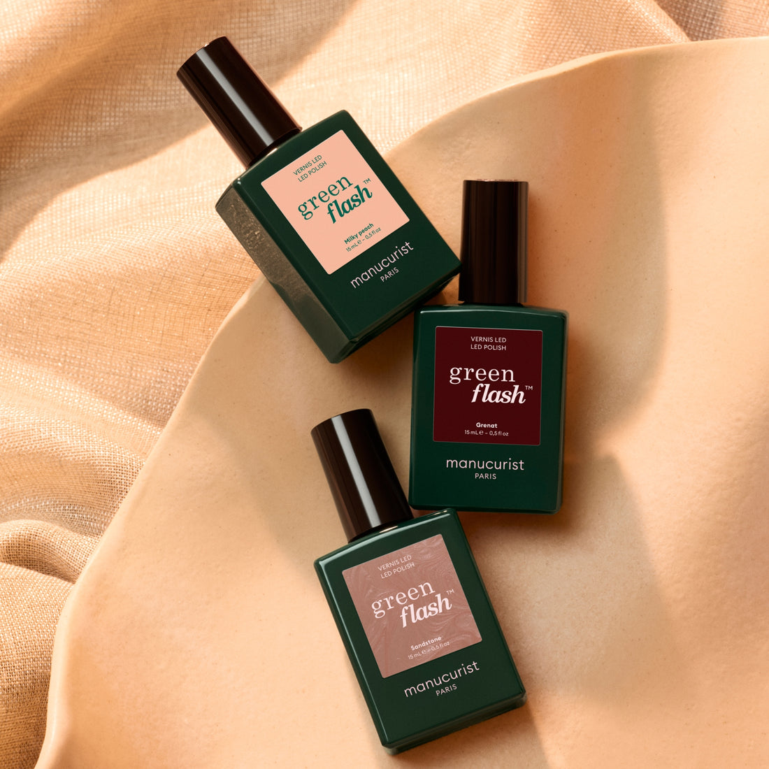 Three bottles from the Manucurist Curated Trios collection rest on a beige fabric. Each bottle, designed for effortless chic manicures, features a distinctive label in blush pink, deep red, and muted teal. Sleek black caps and the minimalist design invite you to discover unique color palettes by Manucurist.