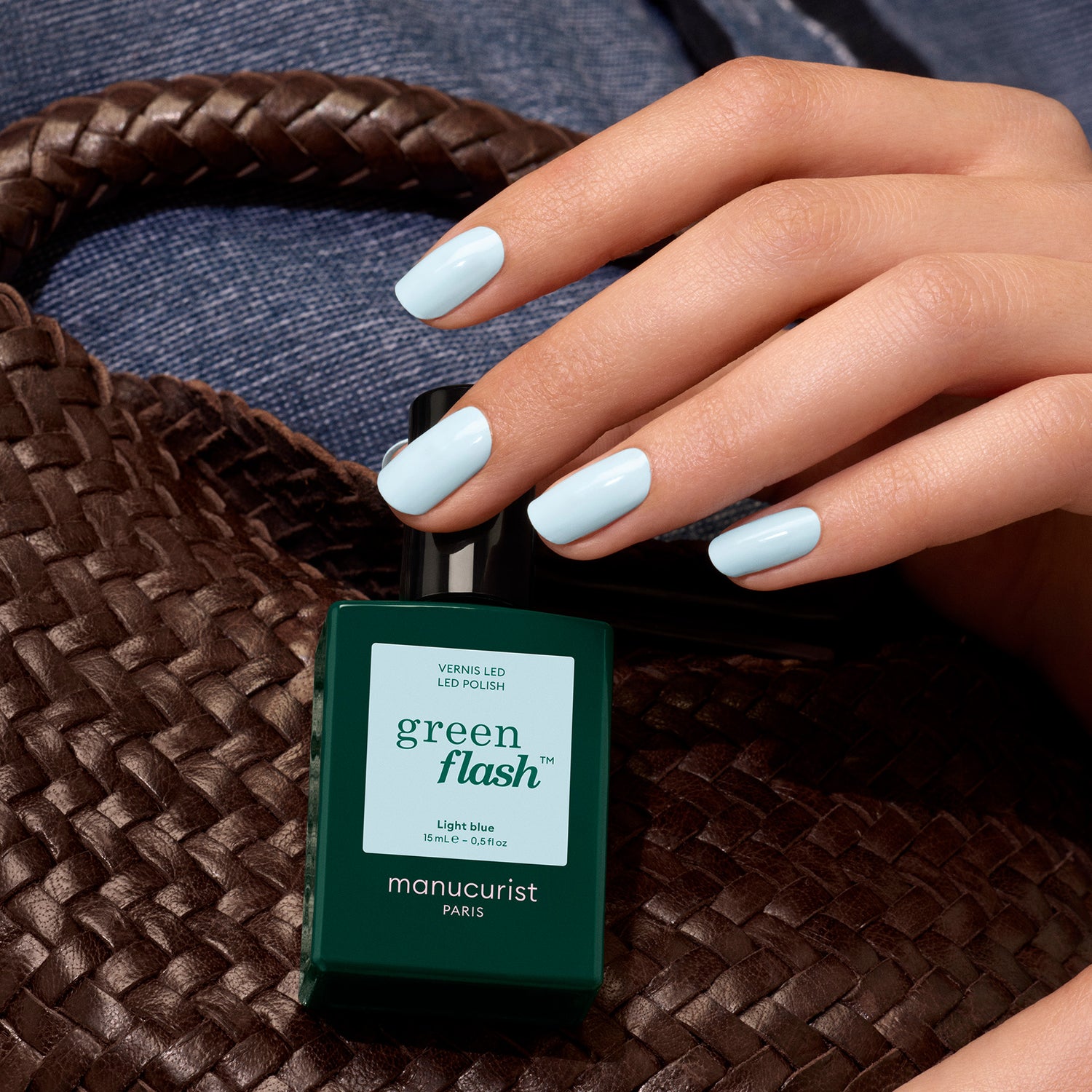 A hand with sky blue gel polish holds a bottle of Manucurist Light Blue polish, with a woven brown bag and denim fabric in the background.