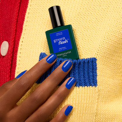 A hand with glossy blue Ultramarine gel polish by Manucurist holds a green Green Flash bottle, tucked into the blue pocket of a yellow and red knit sweater.