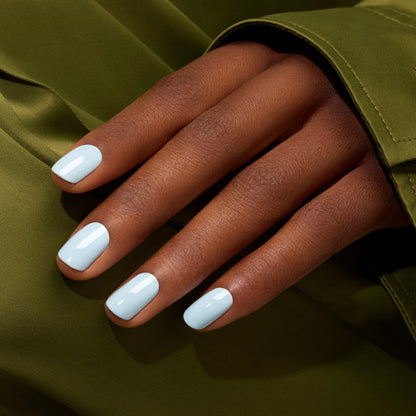 A hand with medium brown skin and neatly manicured nails painted in Manucurist Light Blue nail polish rests on olive green fabric.