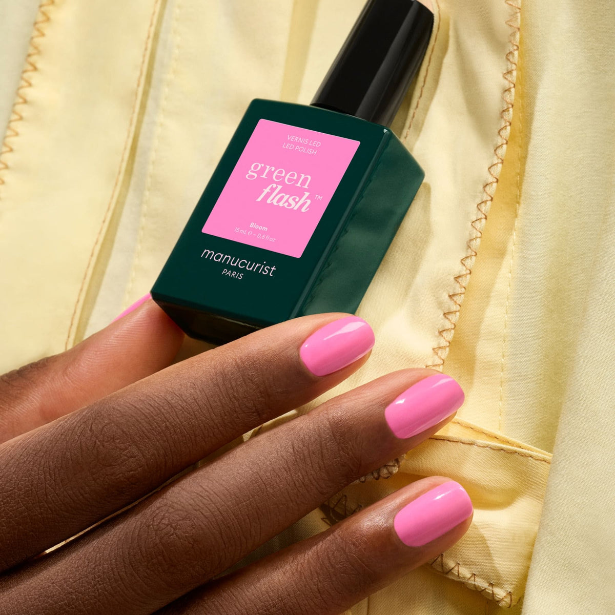 Une main aux ongles roses parfaitement manucurés tient un flacon de vernis gel Bloom de Manucurist, qui se détache sur un fond de tissu jaune.