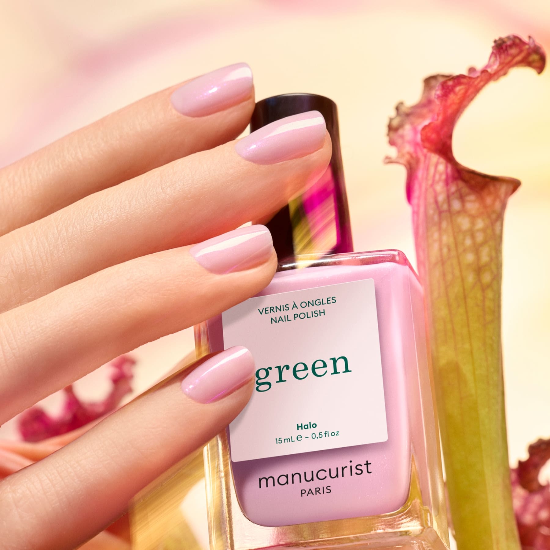 A hand with shiny, light pink milky mauve nails holds a pink "Halo" bottle from Manucurist. A pink flower and green stem are blurred in the background.