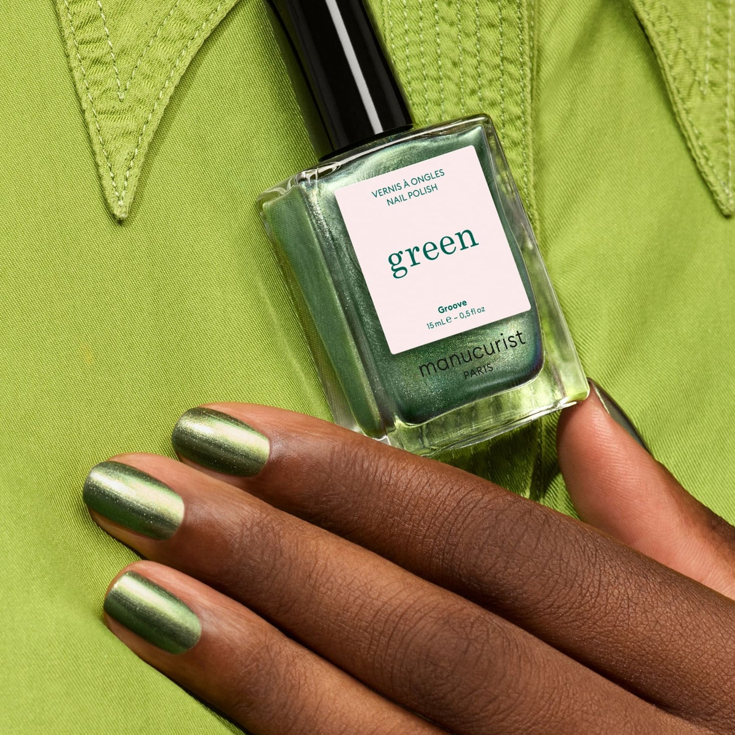 A hand with metallic green nails holds a bottle of Manucurist Groove polish against a lime green fabric background.