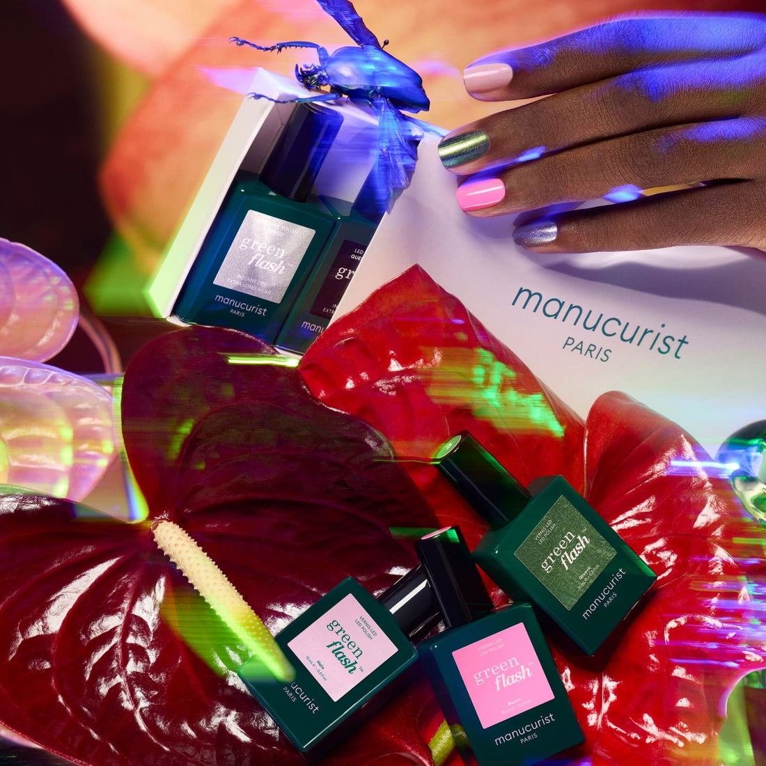 A hand with multicolored nails holds a white Manucurist envelope above Curated Color Collections bottles, vibrant red anthurium flowers, and a glowing insect, all illuminated by bright, colorful lights.
