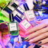 A hand with multicolored nails rests beside five Manucurist Curated Color Collections bottles, surrounded by vibrant flowers and swirling lights, highlighting the latest nail trends and curated palettes for stunning manicures.