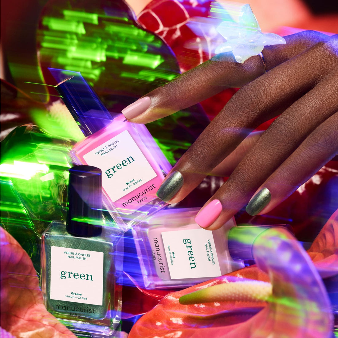 A hand with pink and metallic green nails, inspired by the latest trends, holds two Manucurist Curated Trios bottles among neon-lit plants, with a third bottle nearby and a translucent butterfly ring on one finger.