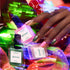 A hand with pink and metallic green nails, inspired by the latest trends, holds two Manucurist Curated Trios bottles among neon-lit plants, with a third bottle nearby and a translucent butterfly ring on one finger.