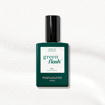 A bottle of Manucurist Bliss gel polish, featuring a dark green label and black cap, is displayed on an iridescent white, subtly sparkling surface.