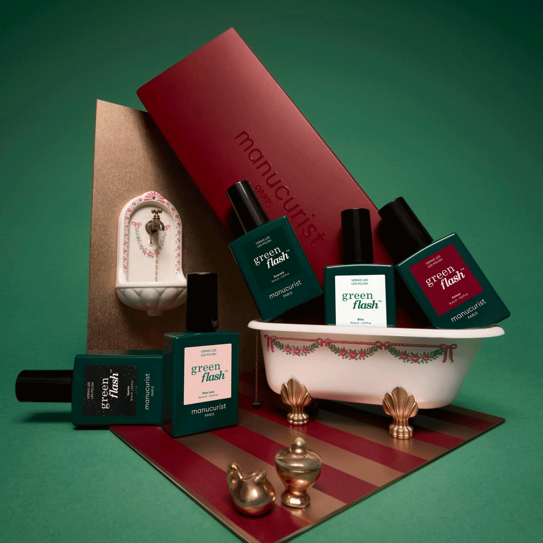 Several bottles from the Manucurist Customizable 5-pack are arranged around a miniature vintage-style bathtub and sink, with a red Manucurist box in the background, all set against a green backdrop.