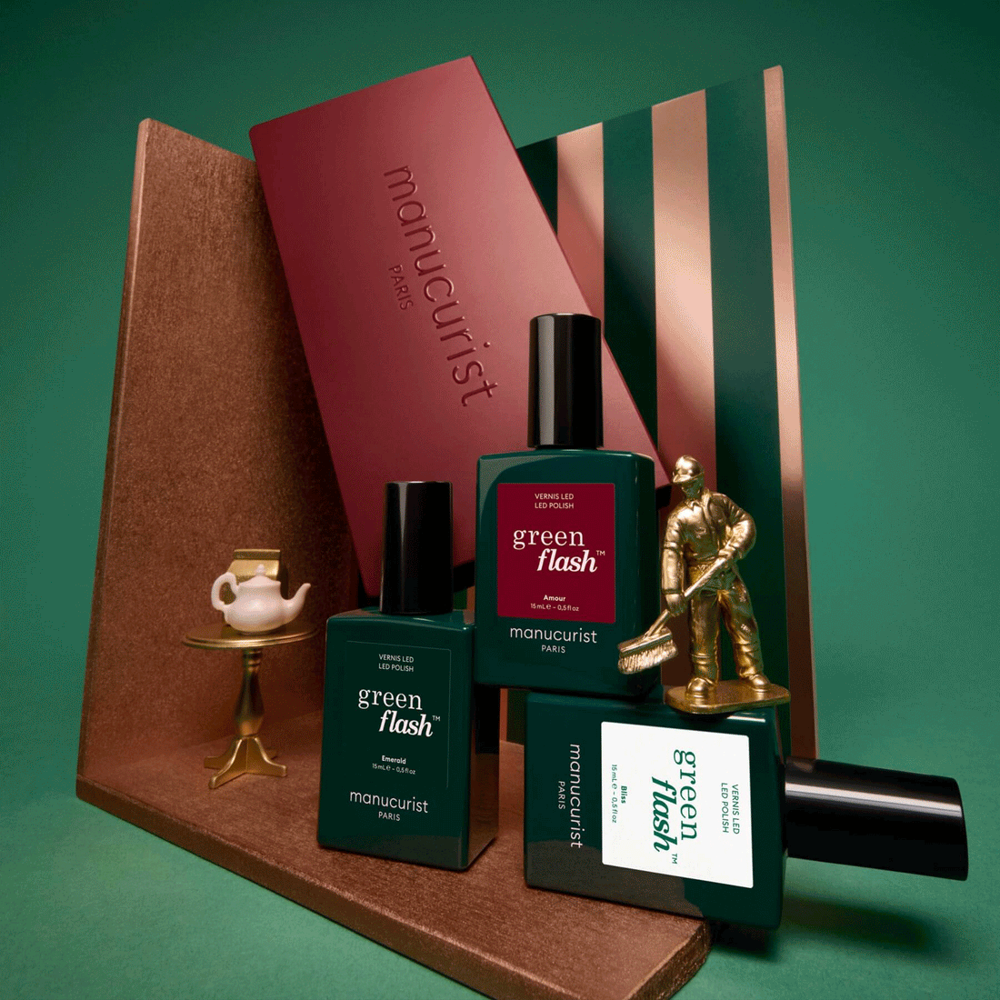 Three green bottles labeled Customizable Trio by Manucurist are displayed alongside a small gold teapot set, a gold statuette, and geometric props against a green background, creating a stylish, modern arrangement.