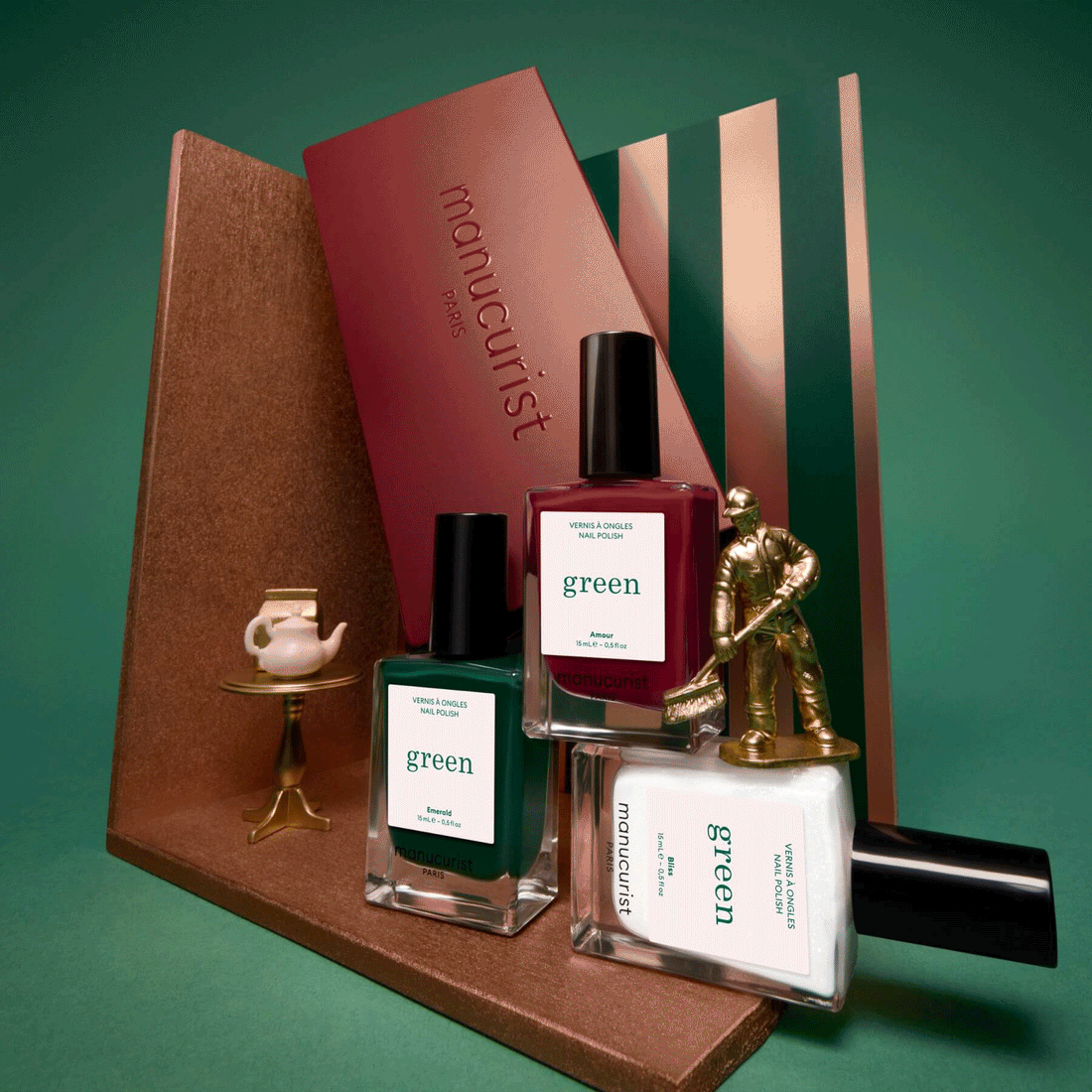 Three bottles of Manucurist’s Customizable Trio—including vibrant green shades—are artfully arranged alongside a gold figurine, a petite gold table with a teapot, and a maroon envelope, all set against a lush green backdrop. This inviting display spotlights the Customizable Trio from Manucurist, inspiring you to assemble your own unique selection for any occasion.