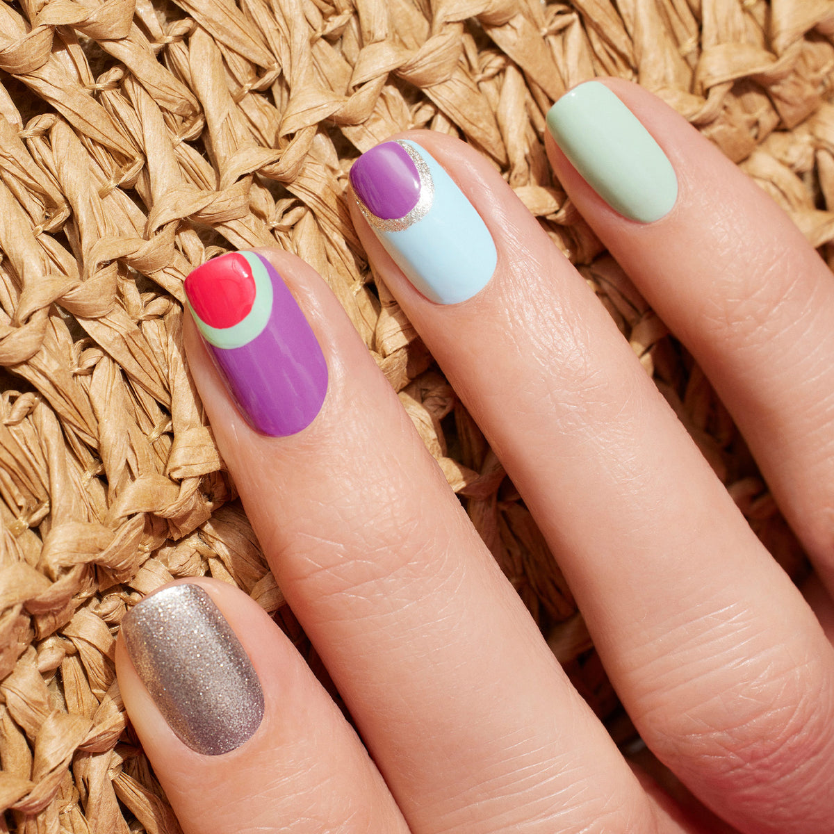 Une main présente les ongles Retro Rainbow de Manucurist : paillettes argentées, violet avec des formes rouges et vertes, bleu avec une pointe violette et une bande argentée, ainsi qu’un vert pastel Green Flash uni — le tout issu de la collection Summer Love — posés sur une surface tissée.