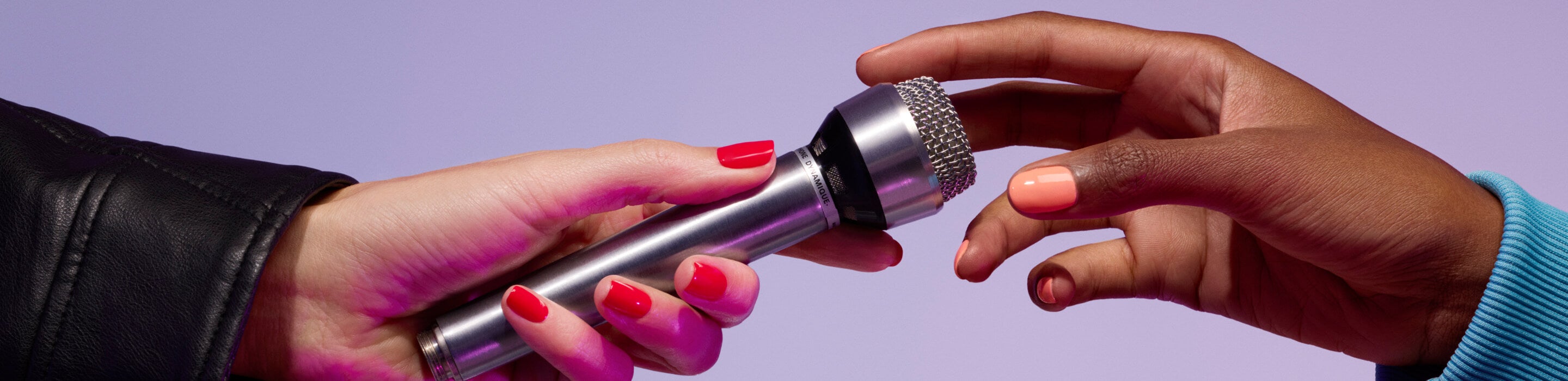 Close-up of two hands exchanging a microphone; one hand has pink nail polish and wears a black sleeve, the other has orange nail polish and wears a blue sleeve, against a purple background.