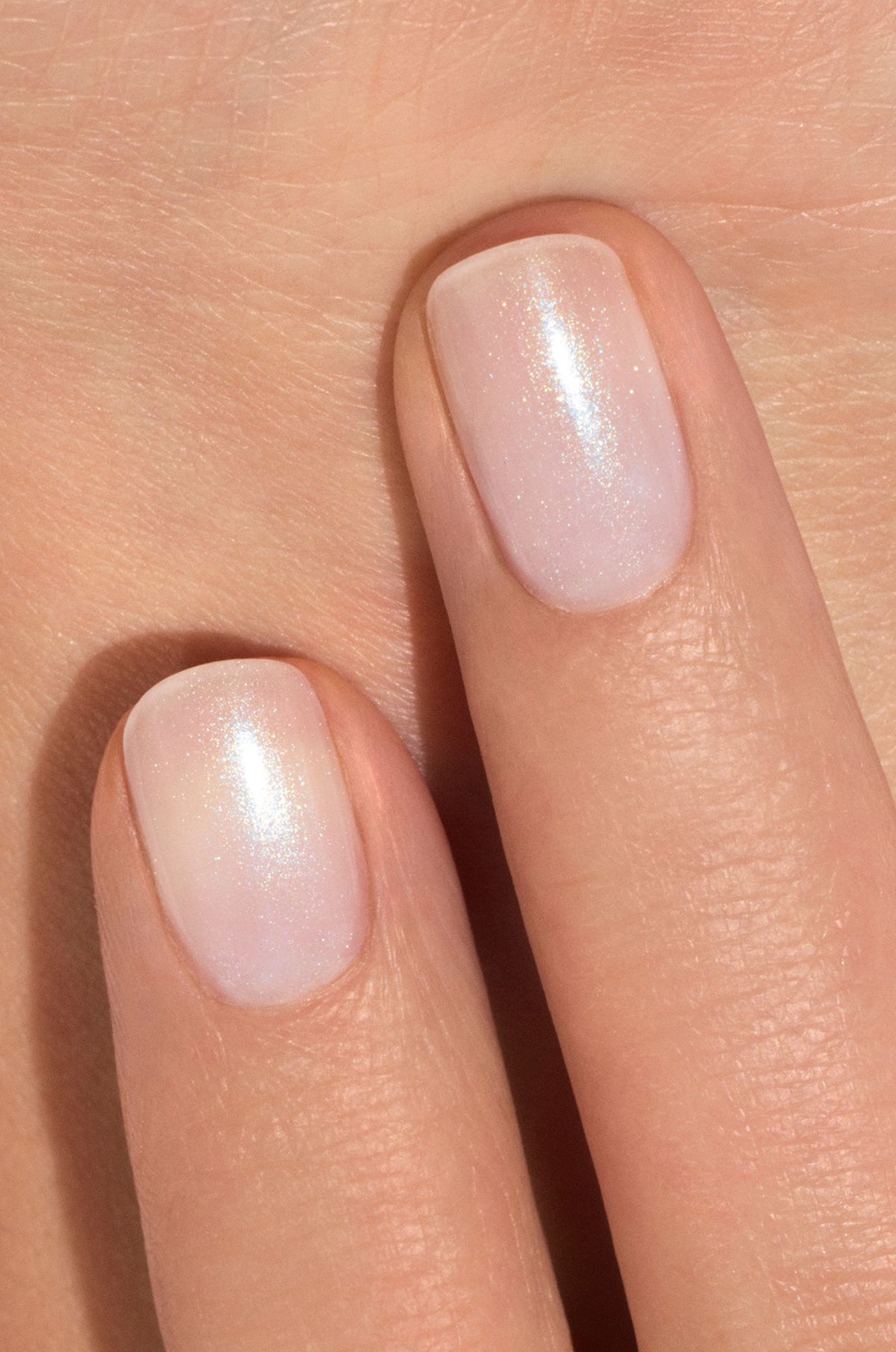 Close-up of two fingers with short, neatly filed nails painted with sheer, pale pink nail polish that has a subtle shimmer, against a light skin background.