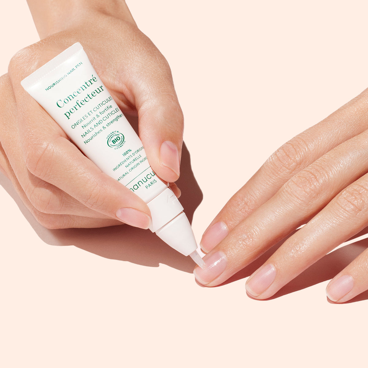 Une main applique le Duo Stronger Nails de Manucurist sur les ongles d’une autre personne, sur un fond rose pâle, pour réparer et perfectionner les ongles et leur donner un aspect sain.