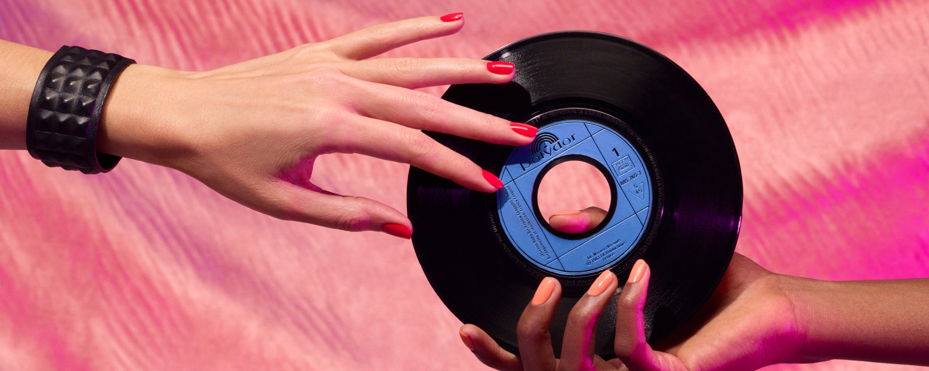 Two hands with painted nails hold a black vinyl record with a blue label against a pink, textured background. One wrist has a black studded bracelet.