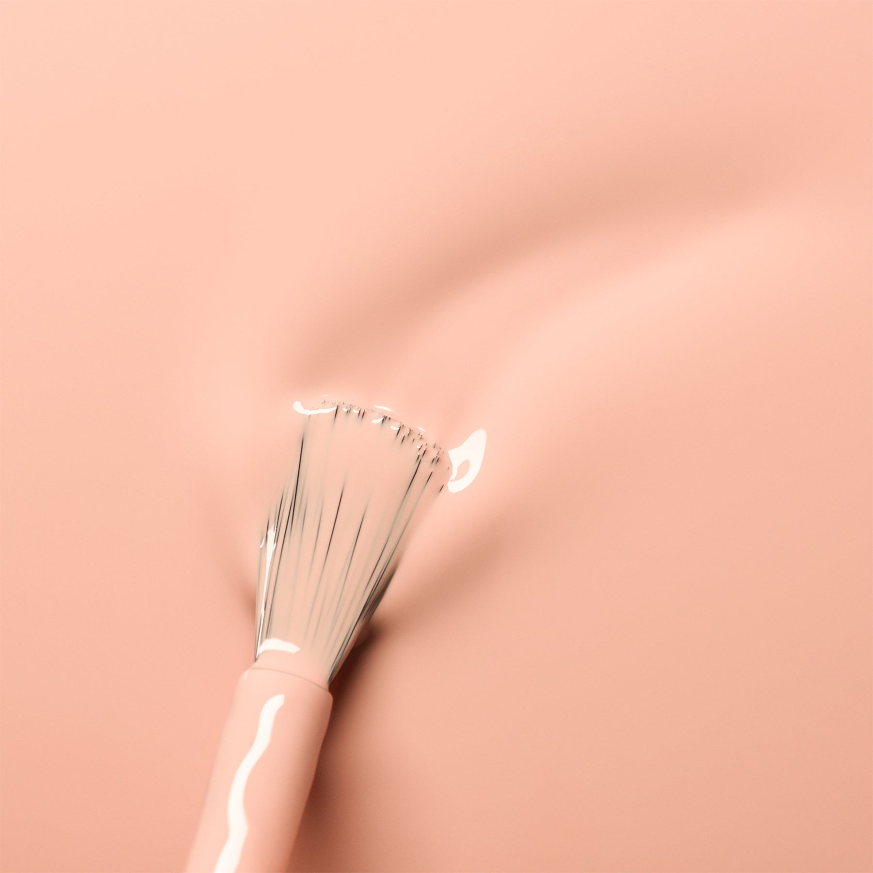 A makeup brush with white bristles swirls on a smooth, peach-pink surface, revealing the soft, glossy Pink Sand shade by Manucurist—a clean, minimalistic look with subtle orange undertones.