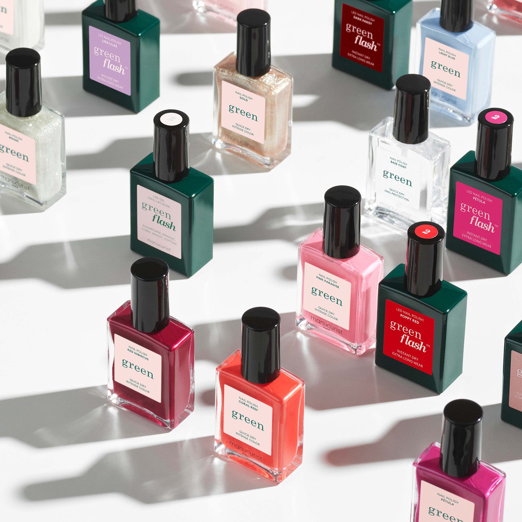 Various bottles of nail polish in different colors are arranged on a white surface, casting soft shadows. Each bottle has a black cap and rectangular shape, with labels reading green or green flash.
