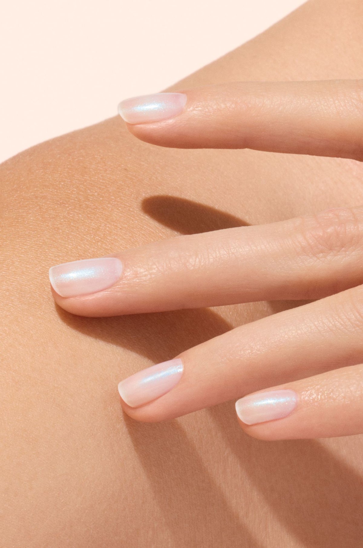 A close-up of a hand with natural, glossy, pale pink manicured nails gently resting on a persons bare shoulder against a soft beige background.