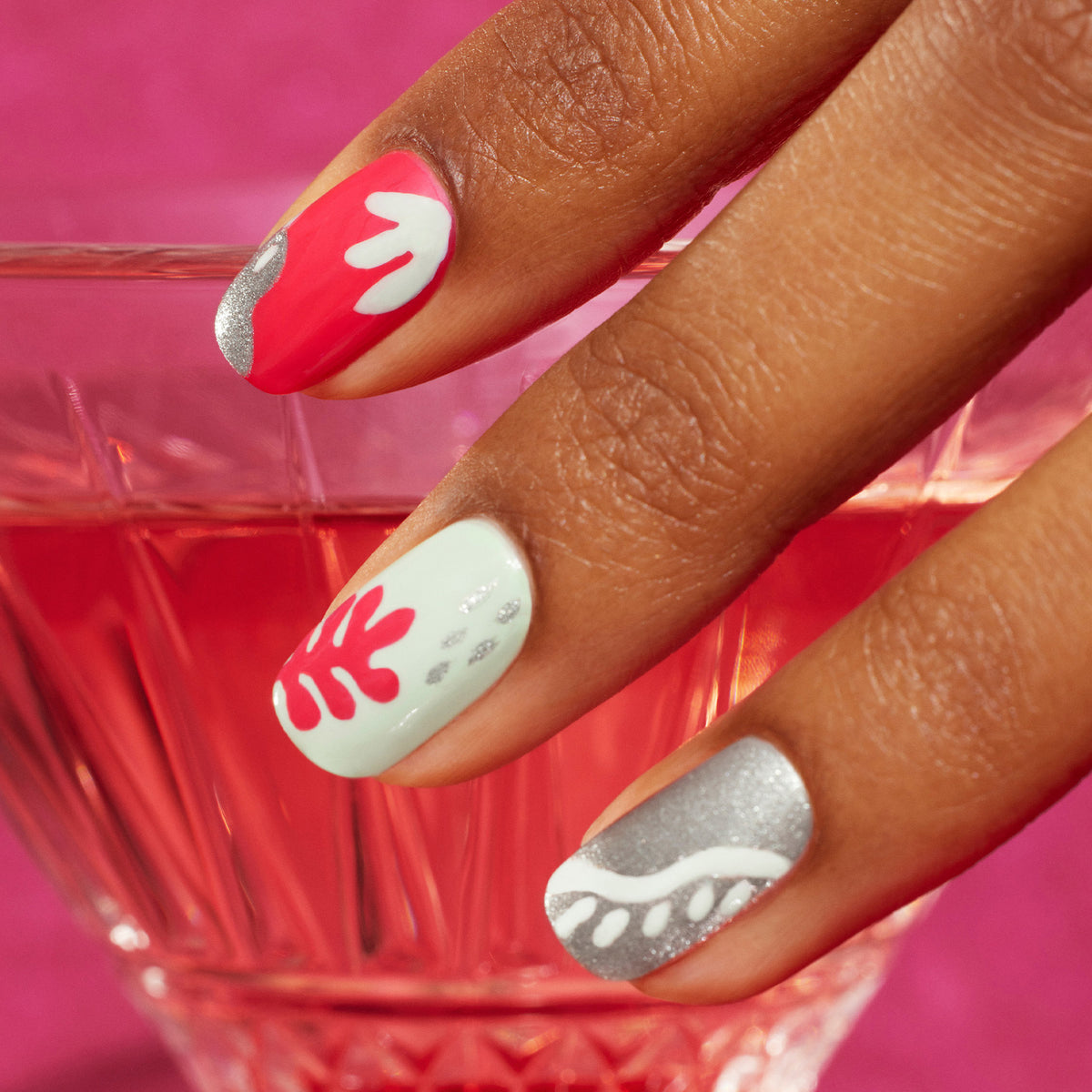 Une main aux ongles manucurés vernis en rouge, blanc, vert menthe et argent avec le Coral Reef de Manucurist, ornés de motifs abstraits ondulés réalisés avec un Nail Art Brush, tenue au-dessus d&