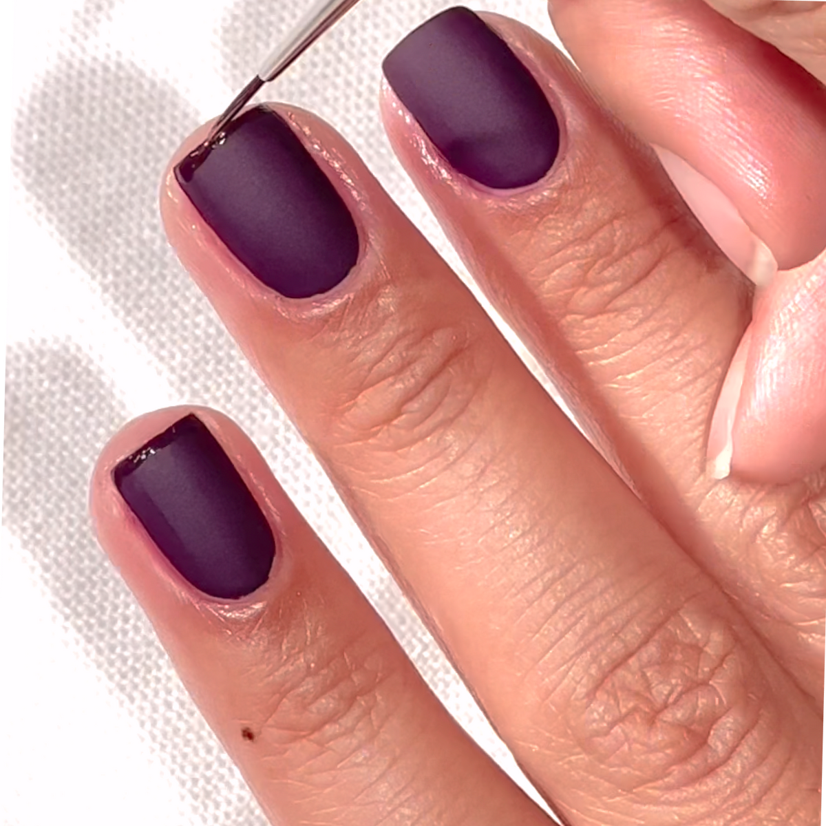 A close-up of a hand with neatly manicured nails painted in a matte dark purple shade, as a thin brush applies polish to the edge of a nail. The background is a white textured surface.